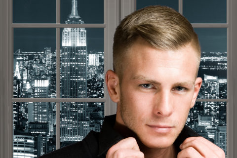 A handsome young blond man stands in front of a window showing New York City at night