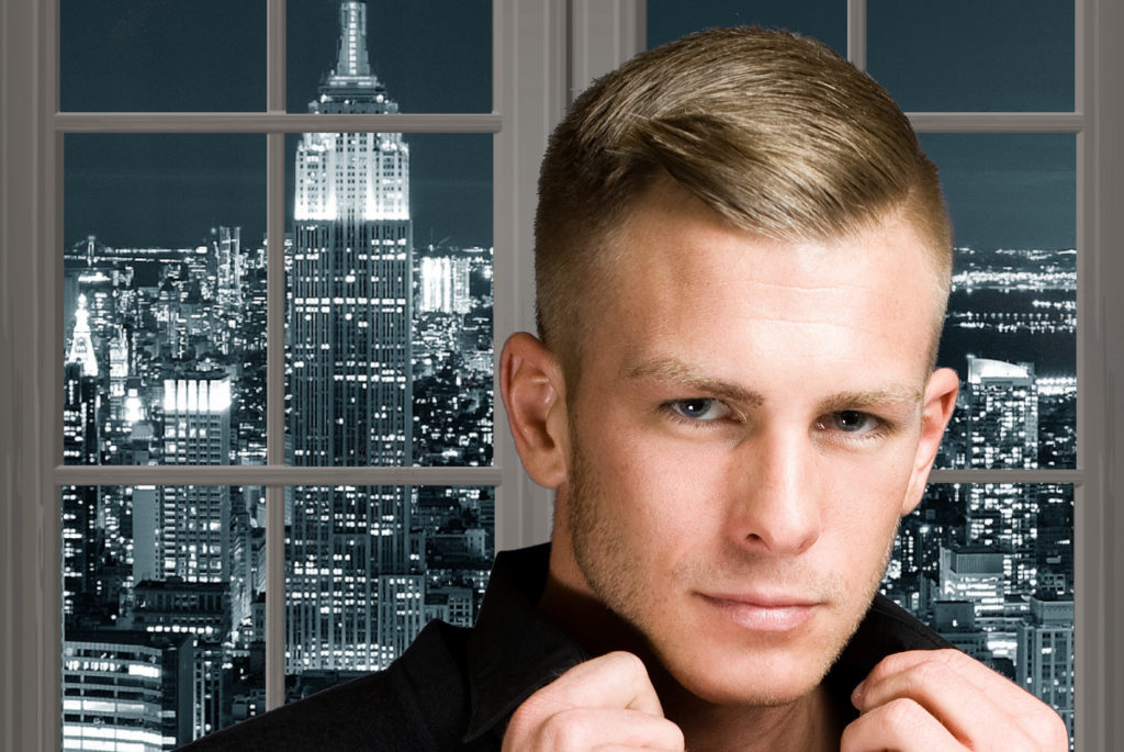 A blond man in front of a window showing a New York City night scene